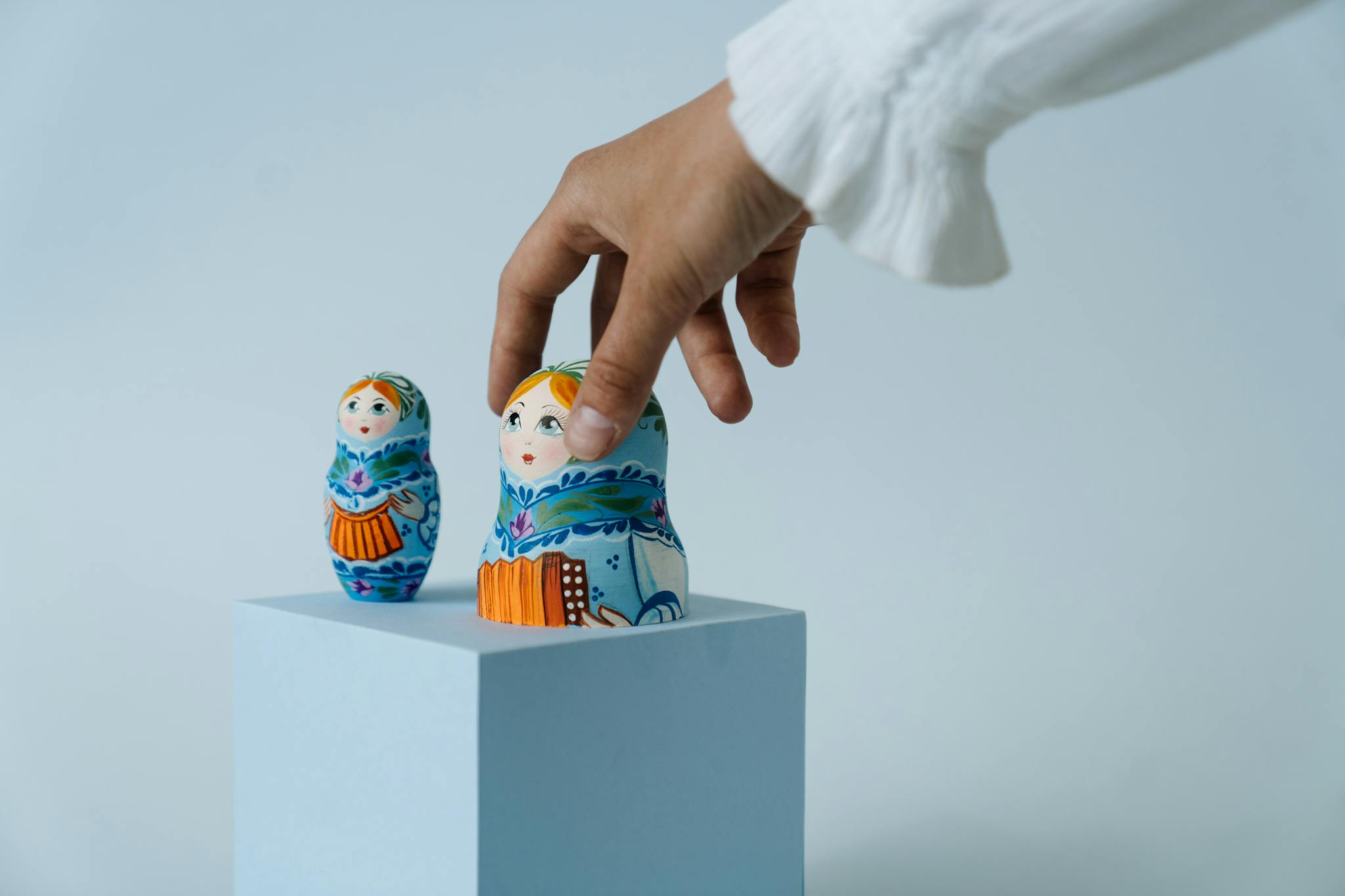 A hand carefully arranging colorful Russian nesting dolls on a blue cube with a soft blue background.