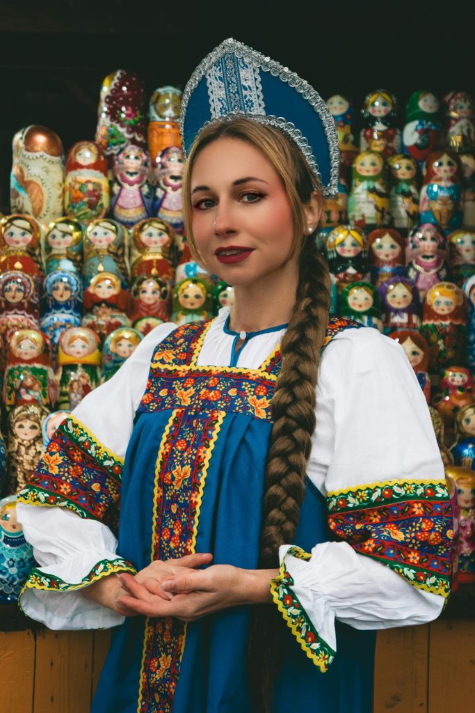 Woman in colorful traditional Russian dress standing in front of Matryoshka doll display.